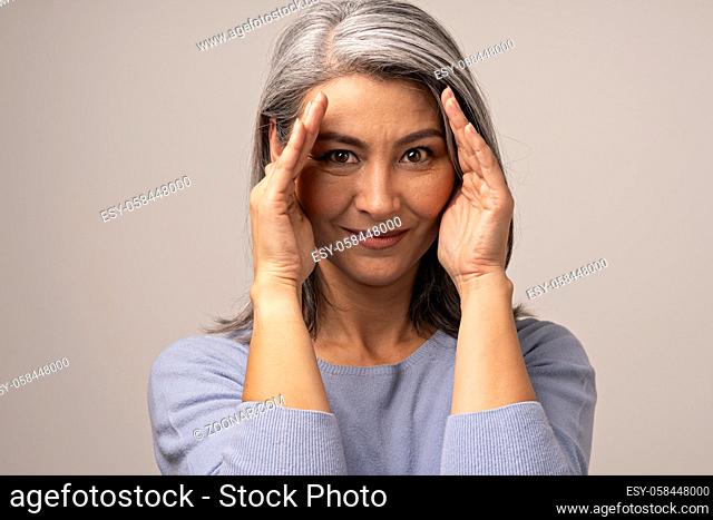 Beautiful Mongolian Woman of Appearance with Gray Hair on a Gray Background. Posing on Camera She Nicely Covers Her Face with Her Hands