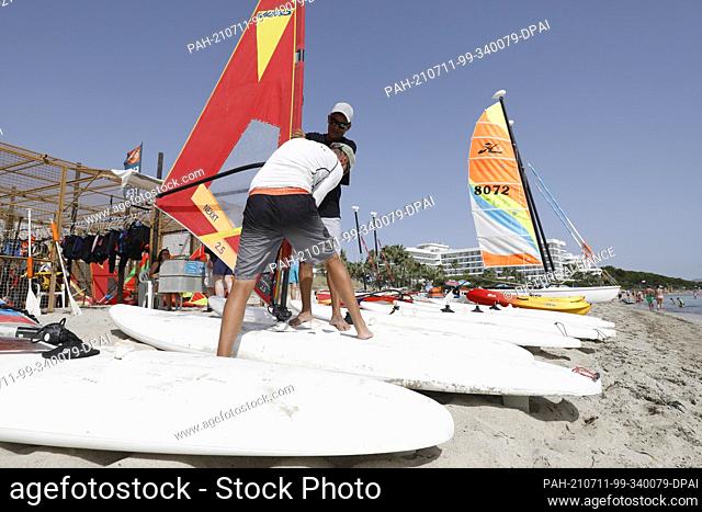 11 July 2021, Spain, -: Teachers of a water sports school set up surfboards for windsurfing on the beach of Playa de Muro in the north of Mallorca
