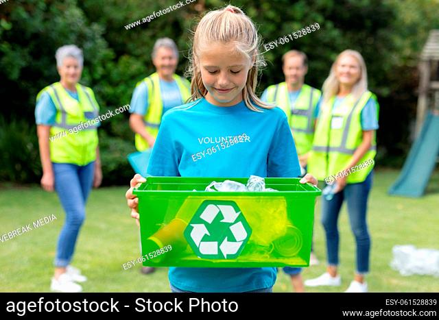 Caucasian girl holding recycling box with group of men and women collecting rubbish in field behind