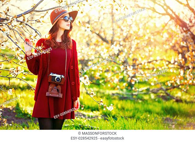 portrait of beautiful young woman with camera and hat standing near blooming tree