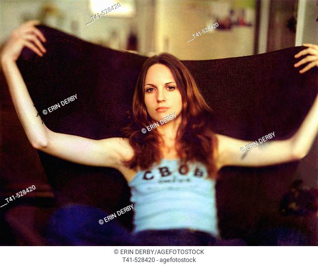 A young woman rests in a brown chair in a small NY apartment. She wears a CBGB's tank top and has many tatoos