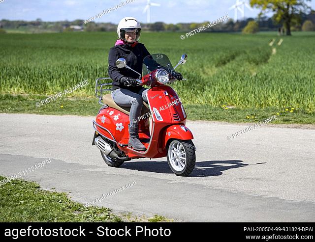 04 May 2020, Schleswig-Holstein, Kiel: In sunny weather, a woman rides her red 125 cc Vespa scooter along a dirt road wearing safety clothing, helmet and gloves