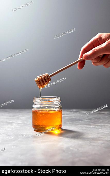 Woman hand hold a wooden stick with dripping flower honey into a glass jar on a gray background, place for text. Pure organic sweet goodness