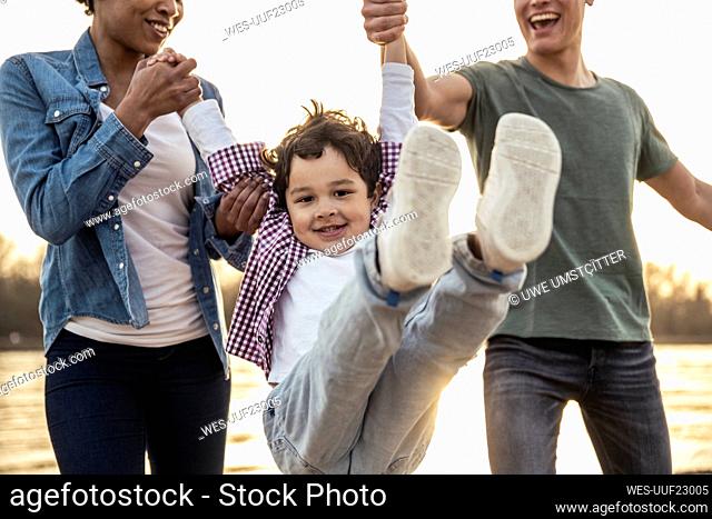 Happy boy swinging while holding hands of parents during sunset