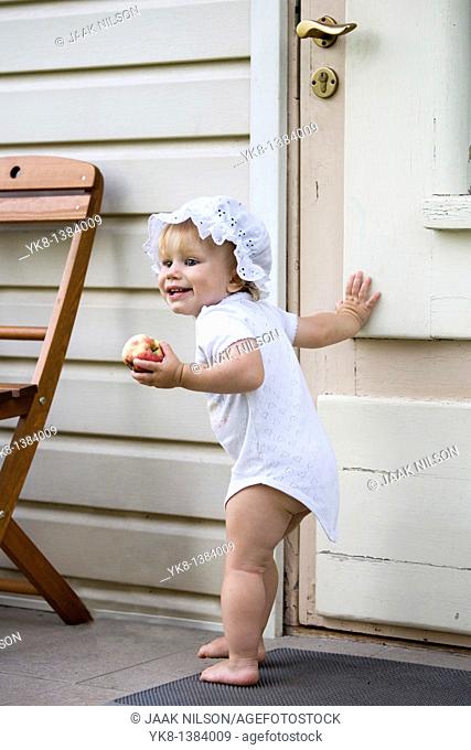 Happy One Year Old Baby Girl Standing Outside by Wooden Door with Apple in Hand