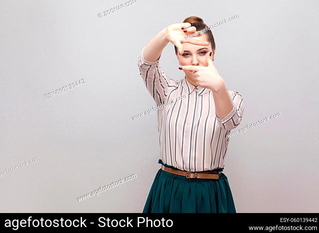 Portrait of beautiful young woman in striped shirt and green skirt with makeup and collected ban hairstyle, standing with cropping composition gesture