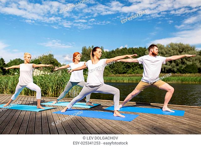 fitness, sport, and healthy lifestyle concept - group of people making yoga in warrior pose on river or lake berth