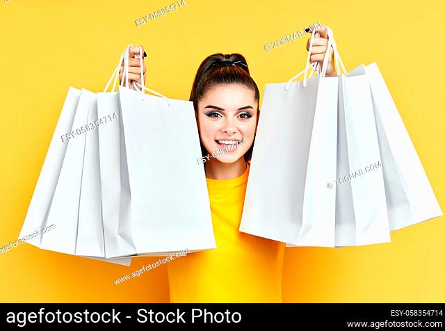 Headshot portrait of young happy excited shopaholic woman with shopping bags in hands enjoy her shopping over yellow background