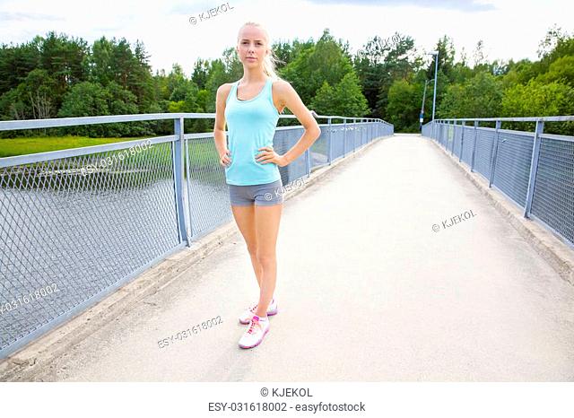Well Trained young blonde woman taking a break after jogging. Standing confident with her hand on her hip on a bridge