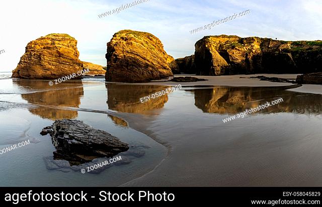 A sandy beach with tidal pools and jagged broken cliffs behind in warm evening light