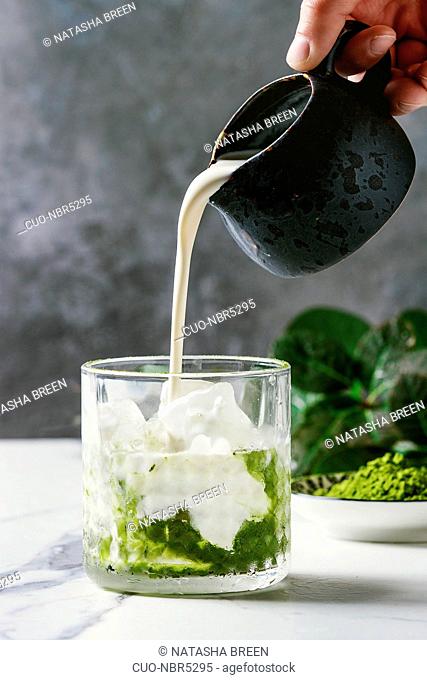Cream pouring from jug to matcha green tea iced latte or cocktail in glass, with ice cubes, matcha powder on white marble table, decorated by green branches