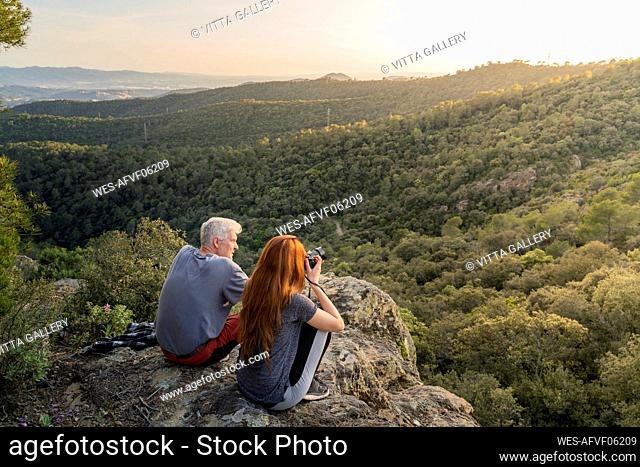 Father and adult daughter sitting on rock enjoying view at sunset