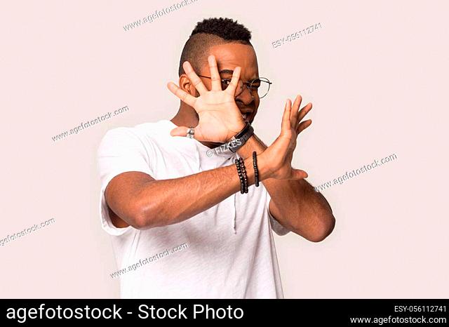 Funny scared African American man in glasses hiding behind hands, watching horror movie, feeling afraid, shy young male refuse gesture, rejecting anything