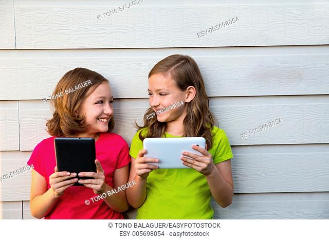 Twin sister girls playing with tablet pc happy on white wall looking each other