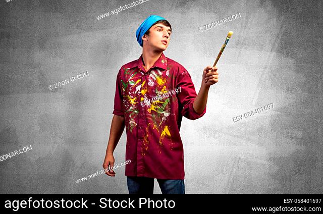 Young emotional artist gesturing with paintbrush. Caucasian painter in dirty shirt and bandana standing on gray wall background