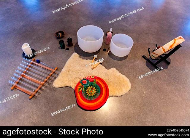 Various sacred objects music instruments like stones, statues, drums, crystal bowls and more are displayed in a set-up on a hard floor, seen from behind