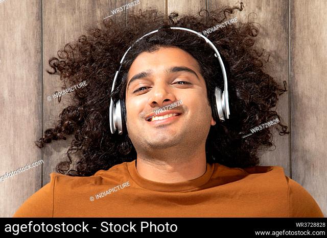 A CURLY HAIRED YOUNG MAN HAPPILY LYING ON WOODEN FLOOR AND LISTENING TO MUSIC