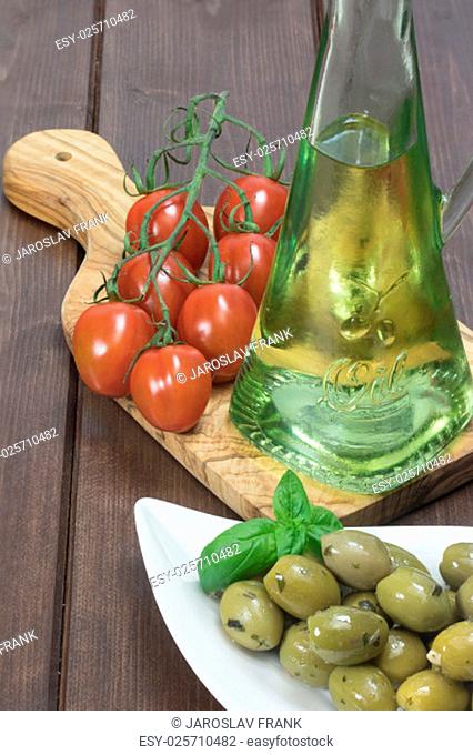 White bowl with marinated green olives is placed on a wooden desk. Tomatoes are lying on a desk of olive wood. Bottle with oil is standing in the background