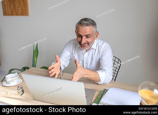 Ability to convince. Emotional gray-haired man talking on video call holding hands at level of laptop screen sitting at table