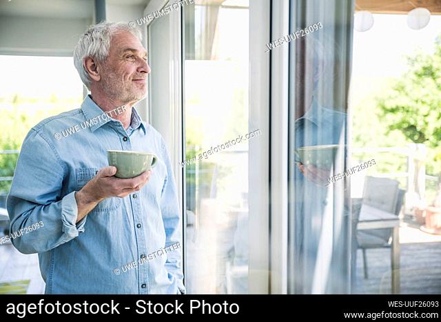 Happy senior man holding coffee cup looking out through window