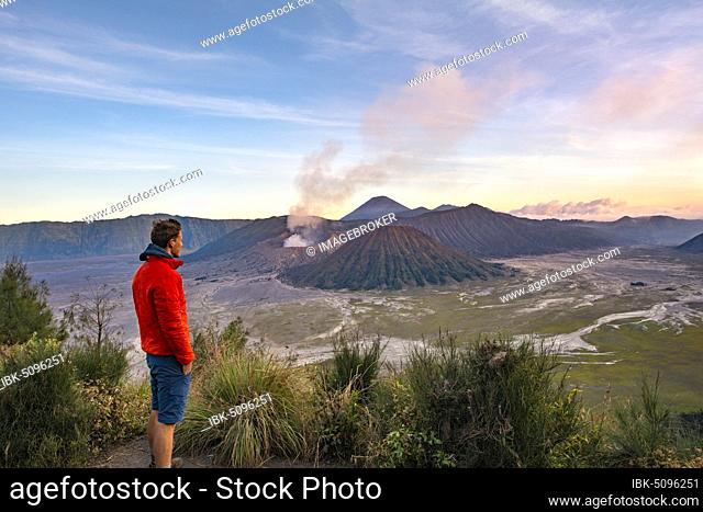 Young man in front of volcanic landscape at sunset, view in Tengger Caldera, smoking volcano Gunung Bromo, in front Mt. Batok, in the back Mt
