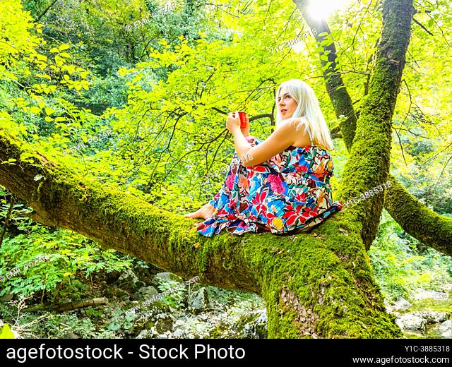Attractive blonde woman is siting on a tree with a tea-pot in hands looking away