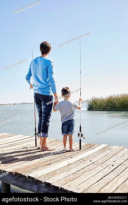 Father and son with fishing rods holding hands while standing on pier