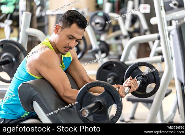 Young handsome man exercising bicep curls with the E-Z barbell while sitting down at preacher curl bench during upper-body workout routine