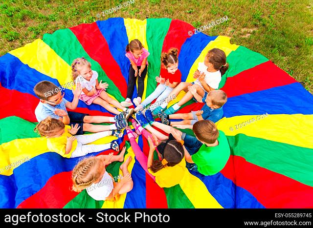Kids play outdoor, sitting on rainbow parashute, clap their hands