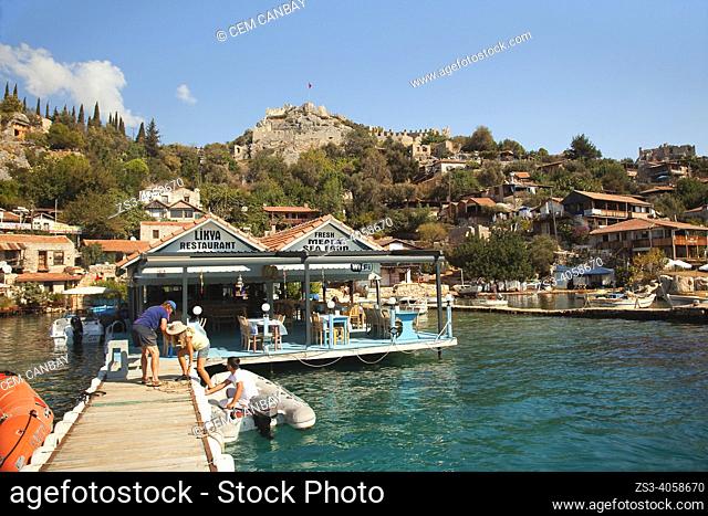 View of the Simena Castle with the tourists climbing up a pier in front of the houses and local restaurants in Kalekoy, Ucagiz village, Demre, Antalya Province