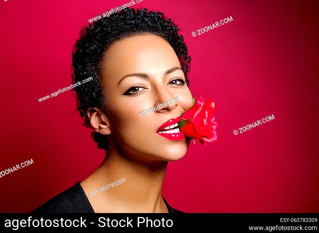 beautiful young woman with red lips holding rose in mouth. studio beauty shot on dark red background. copy space