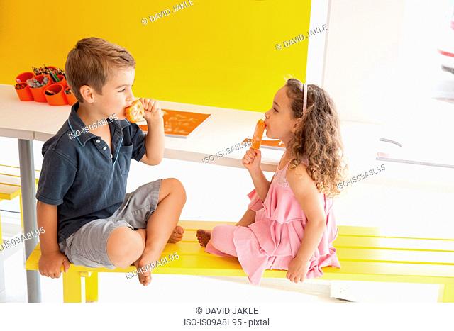 Brother and sister eating ice lollies on bench indoors