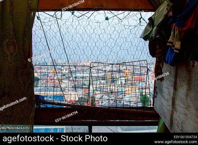 View of the city with colorful houses and commercial buildings from a shabby window with a metallic net protection and wood planks worn blinds with clothes