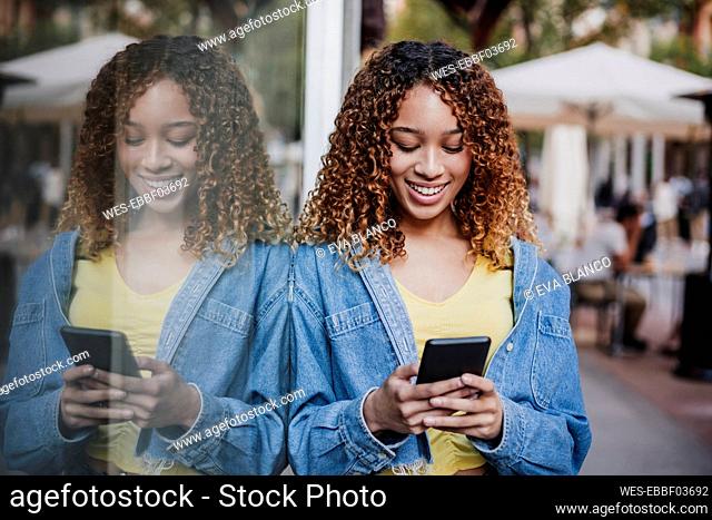 Young woman using smart phone while leaning on glass wall