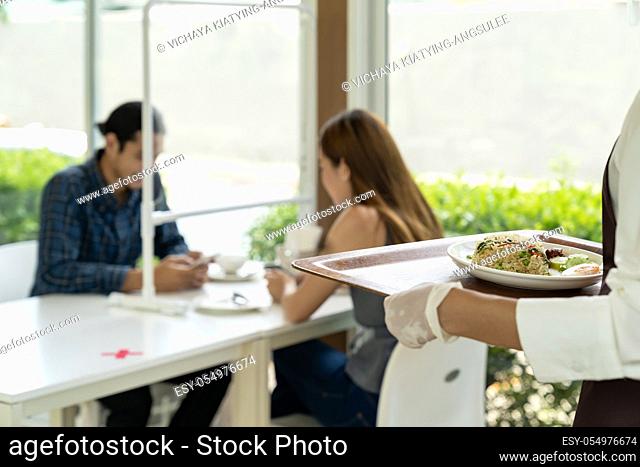 back view asian waitress holding food tray to serving meal to customer with customer in background. New normal restaurant lifestyle concept.