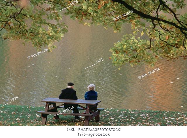 Seniors on a park bench enjoy fall colors reflected in the water, Ontario, Canada  Early autumn, Maple tree overhead will shed all leaves in a week or two