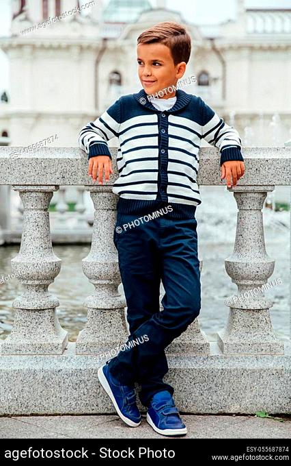 Stylish rich little boy in fashionable clothes. Leaned on Granite-stone handrails. stylish, handsome posing outside in the city park
