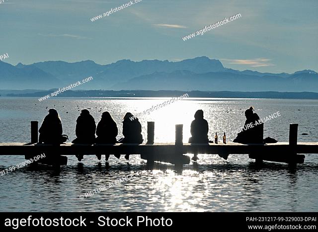 17 December 2023, Bavaria, Starnberg: Excursionists sit on a jetty on Lake Starnberg and enjoy the sun and the view of the Zugspitze