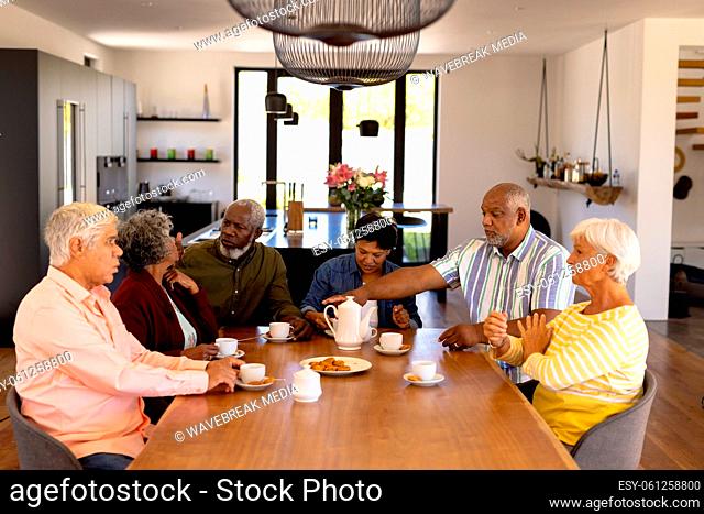 Senior multiracial friends talking and enjoying coffee with cookies at dining table in nursing home