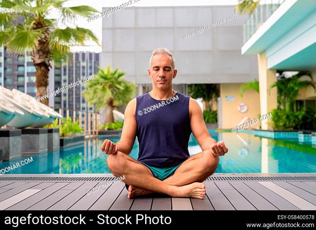 Portrait of mature handsome Persian man with gray hair relaxing at the swimming pool on rooftop