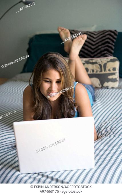 Barefoot latina teen girl uses laptop computer while lying on a bed