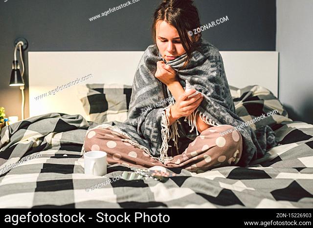 The theme is seasonal cold, runny nose flu virus infection. Young Caucasian woman at home bedroom bed sitting wrapping blanket, chill heat high temperature