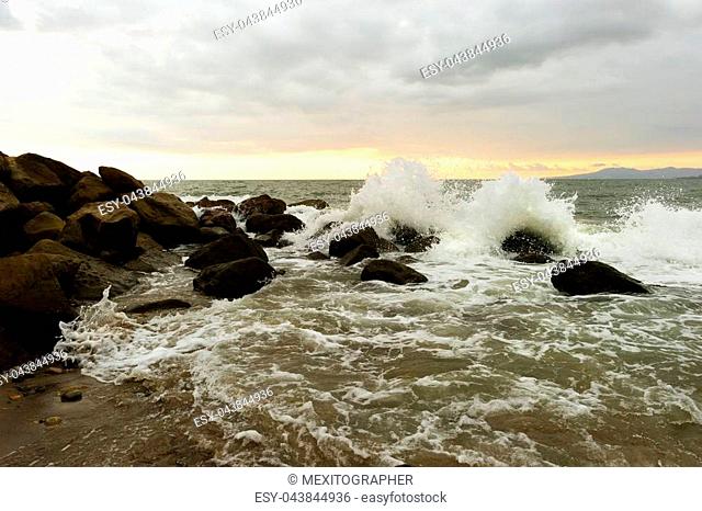 Waves crashing is an ocean wave rushing to shore hitting sea rocks and shooting up into the air as ocean spray