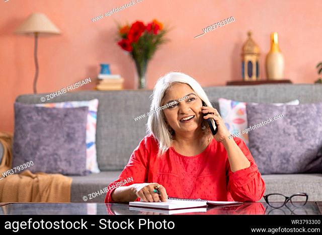 Old woman talking on mobile phone while writing on notepad at home