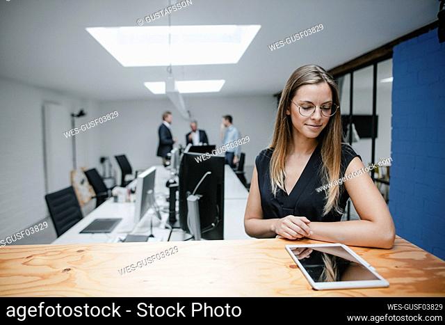 Businesswoman standing in office, leaning on wooden counter, using digital tablet
