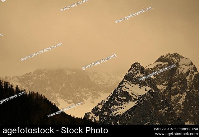 15 March 2022, Bavaria, Krün: Over the peaks of the Wetterstein mountains bad weather with Sahara dust is coming up that colors the sky yellow/orange