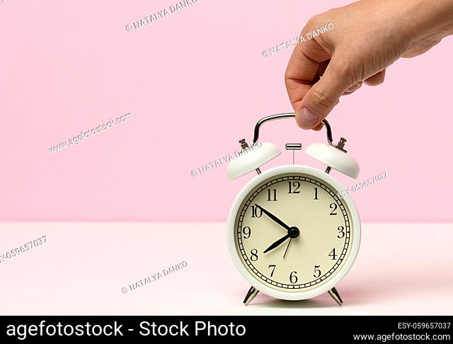 female hand holds a white metal alarm clock on a white table, time is ten to eight in the morning