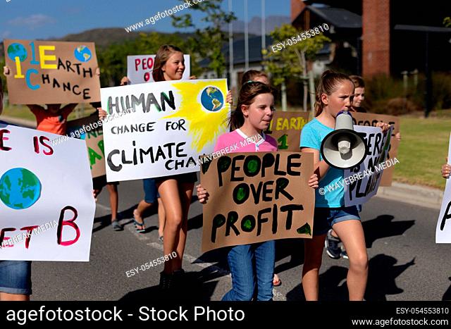 Front view of a diverse group of elementary school pupils walking down a road in the sun on a protest march, carrying signs with environmental and conservation...