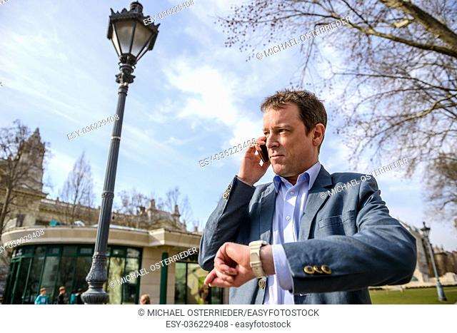 A middle age businessman standing in a park while talking on his phone and checking the time on his watch