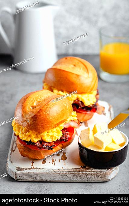 Breakfast sandwiches with scrambled egg, bacon, cheese, tomato on white wooden board, glass with fresh orange juice, white background